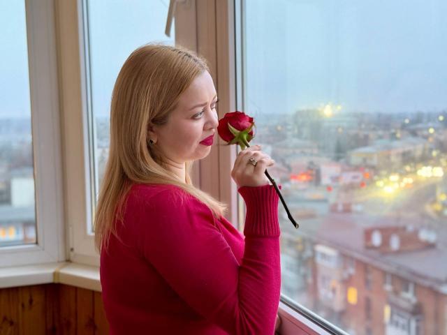 OliviaVerr steht vor einem Fenster und hält eine rote Rose an ihre Nase, sie trägt einen roten Pullover. Ihre blonden Haare sind glatt, und sie trägt roten Lippenstift. Im Hintergrund ist eine Stadtansicht zu sehen..