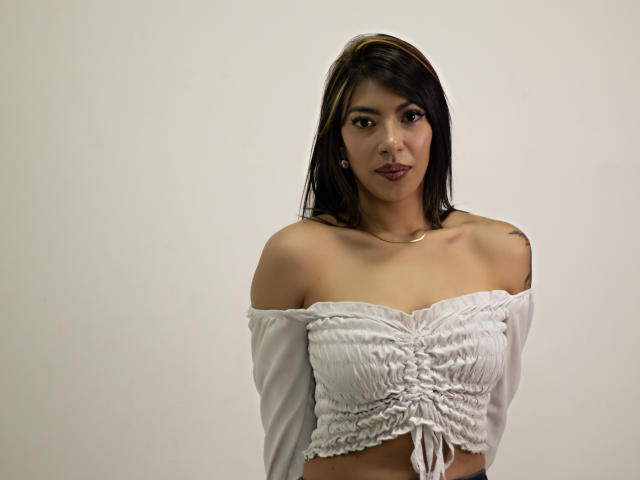 Fotografie von ZoediMaria, einer attraktiven Frau mit mittelbrauner Haut und schwarzen Haaren, die ein weißes Top mit einem Schulterfrei-Schnitt trägt. ihr Dekolleté zur Schau stellend, direkt in die Kamera blickend mit einem verführerischen Ausdruck.