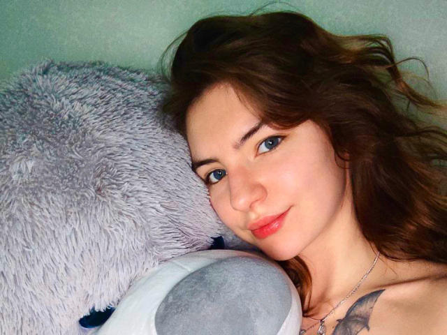 AlisaBell69 está deitada contra um animal de pelúcia cinza e fofo, com a cabeça ligeiramente inclinada, olhos azuis, lábios vermelhos, pele clara e cabelo preto. cabelo, pequena tatuagem no ombro, casual, sexy, relaxada, íntima.