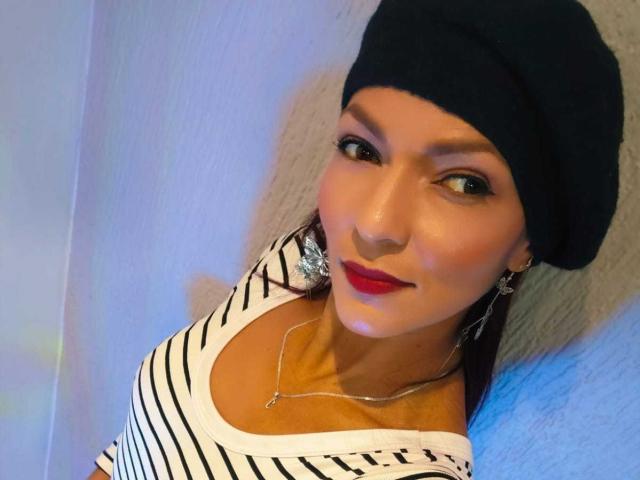 Foto ravvicinata di HannaPretty, che indossa un berretto nero e una maglietta a righe bianche, con rossetto rosso e orecchini argentati. Sta guardando la fotocamera con un leggero sorriso, con una luce blu sullo sfondo..