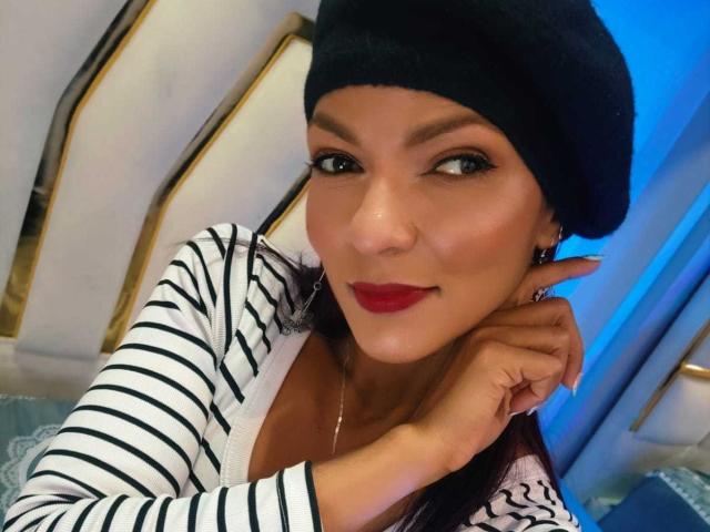Close-up of HannaPretty with olive skin, black beanie, red lipstick, and striped shirt, hand resting on neck, seductive gaze, blue light in background, slight smirk, intense sexual tension.