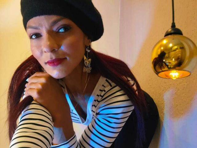 HannaPretty, brown-skinned woman with long red hair, black beanie, striped shirt, large earrings, blue lighting, gold pendant lamp, slightly smirking, hand near chin, soft light, sexual tension.