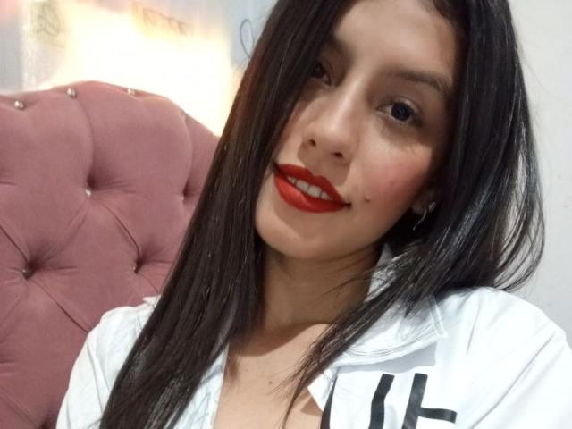 Close-up of MadissonFontaine with long black hair, red lipstick, and white shirt, leaning against a pink tufted chair, looking directly at the camera with a slightly seductive expression.