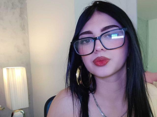 Photograph of IsabellaaRossi, a curvy woman with pale skin and black hair, wearing glasses, red lipstick, and hoop earrings, looking directly at the camera, with a lamp in the background.