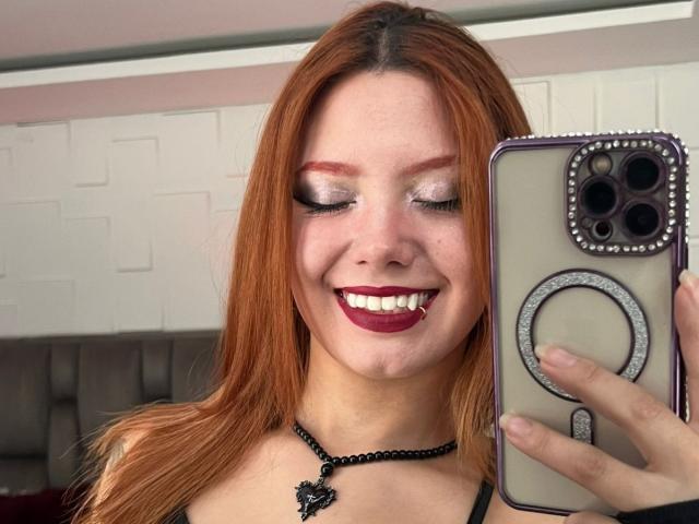 Close-up photo of EmmaThomson with long red hair, smiling with red lipstick, holding a phone with a circular ring case, black necklace with a pendant, white brick wall background. Fucking selfie.