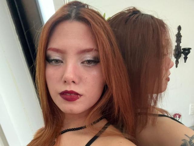 Close-up van EmmaThomsonn met lang rood haar, veel make-up, donkerrode lippenstift en een zwarte choker. Ze staat voor een spiegel, die haar reflecteert, en waarop een tatoeage op haar rechter schouder te zien is. Heel aantrekkelijk..
