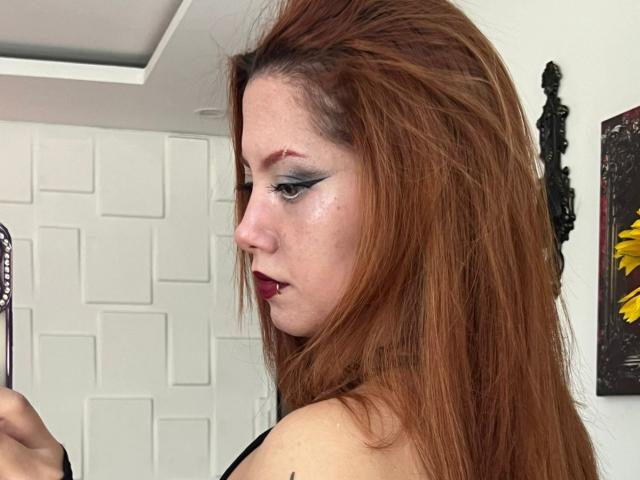 Close-up photo of EmmaThomson with long, red hair, heavy black eyeliner, and dark lipstick. She's looking down with a slightly serious expression. Background includes white tiles and black art.