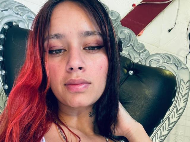 Foto em close-up de RoxanaSanchezz, com pele clara e cabelo ruivo-preto, olhos semicerrados, lábios ligeiramente entreabertos, vestindo uma colar, apoiado em uma cadeira de couro preto com uma moldura prateada e ornamentada. Muito atraente..