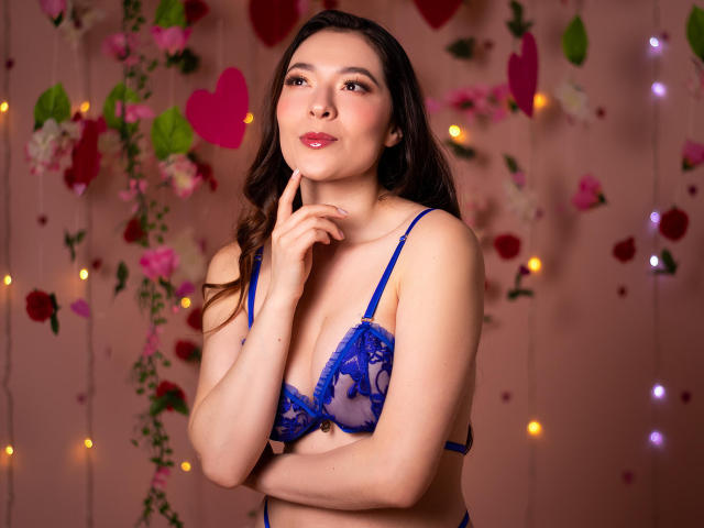 TaniaSandersX, con cabello castaño largo, vestida con un sostén de encaje azul, posa con un dedo en el mentón. Fondo: pared rosa, flores de colores e luces de hadas brillantes. Expresión seductora y segura..