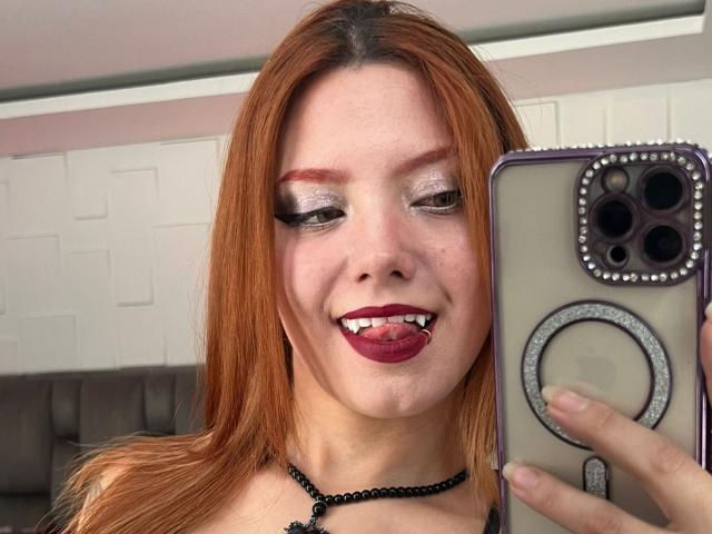 Photo en gros plan d'EmmaThomsonn avec des cheveux roux, un maquillage prononcé et un rouge à lèvres foncé, tirant la langue et tenant un téléphone. Boîtier de bague, collier ras du cou noir, peau claire, fond clair, légèrement flou..