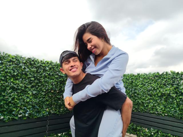SalomeAndAustin, a dark-haired woman in a light blue shirt, hugs a young man with a black cap and black shirt, smiling at each other. Green hedge background, black bench.