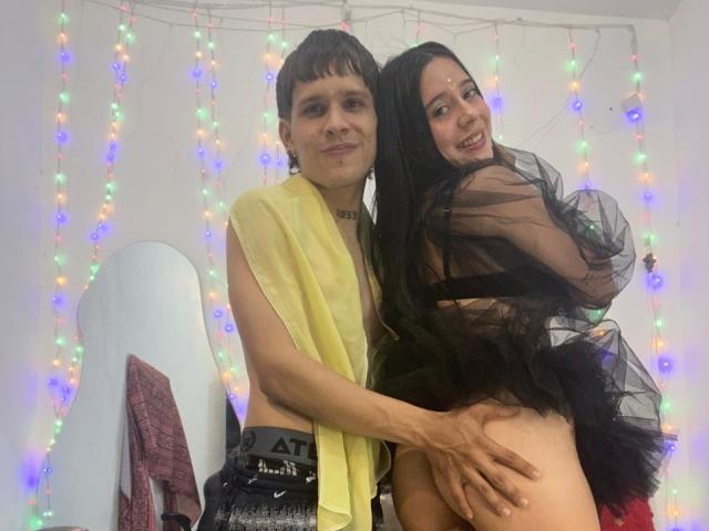 AnjaliRohan, com cabelo curto e escuro e um lenço amarelo, agarra o traseiro nu e redondo de outra AnjaliRohan com cabelo longo e preto. cabelo e uma roupa preta transparente, luzes coloridas em segundo plano.