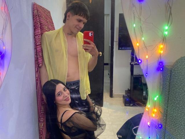 AnjaliRohan, sin sostén, vistiendo una camisa amarilla transparente, sosteniendo un teléfono, está de pie detrás de una mujer que está de rodillas y tiene el pelo largo y negro. con una blusa negra transparente, rodeada de luces navideñas de colores.