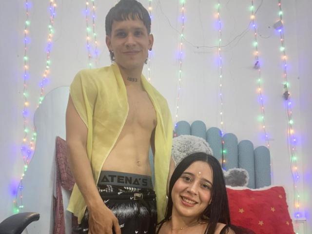 AnjaliRohan with short brown hair stands behind a smiling woman with long black hair, wearing a yellow shirt, exposing his chest and black "Aetna" pants. String lights and pillows in background.