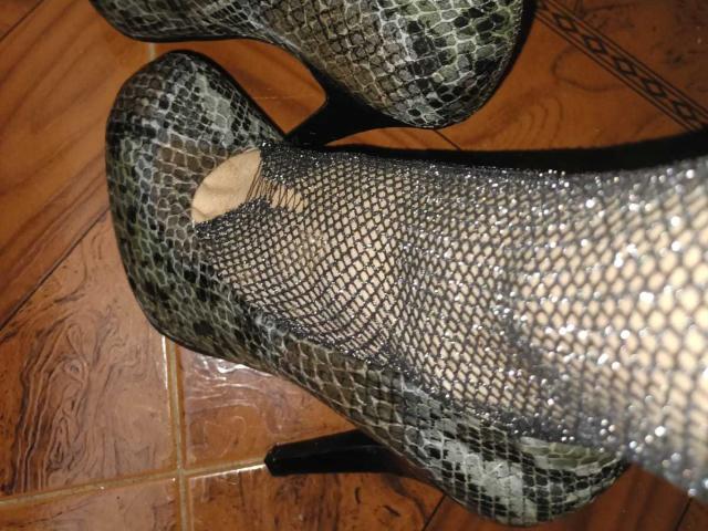 Nahaufnahme eines High-Heel-Schuhs mit Schlangenmuster, in Schwarz und Grau, mit kleinen, offenen Rissen, die einen ein fleischfarbener Fleck auf einem Holzboden. Glänzende, strukturierte Oberfläche.