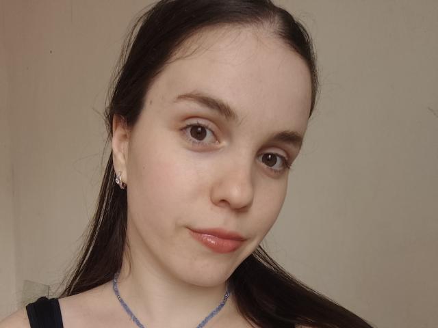 Primer plano de Olyanash, con piel clara, ojos marrón oscuro y cabello negro lacio, vistiendo una blusa negra y un collar azul. Sonrisa suave, maquillaje mínimo, un ligero rubor. El fondo es beige liso..