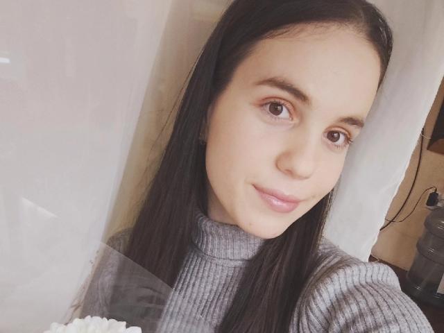 Fotografía de primer plano de Olyanash, con piel pálida, cabello negro largo y ojos marrones, vistiendo un cuello alto gris. Ella mira directamente a la cámara con una sonrisa sutil y sensual. Muy atractivo/a..