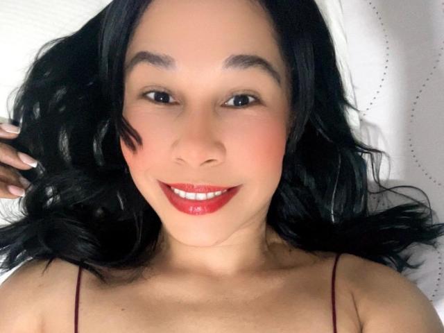 Close-up foto van IvannaX69 met halflang zwart haar, rode lippenstift en een lichte glimlach. Ze heeft een lichte huid, donkere ogen en draagt een top met dunne bandjes. Witte achtergrond. Duidelijke seksuele spanning..