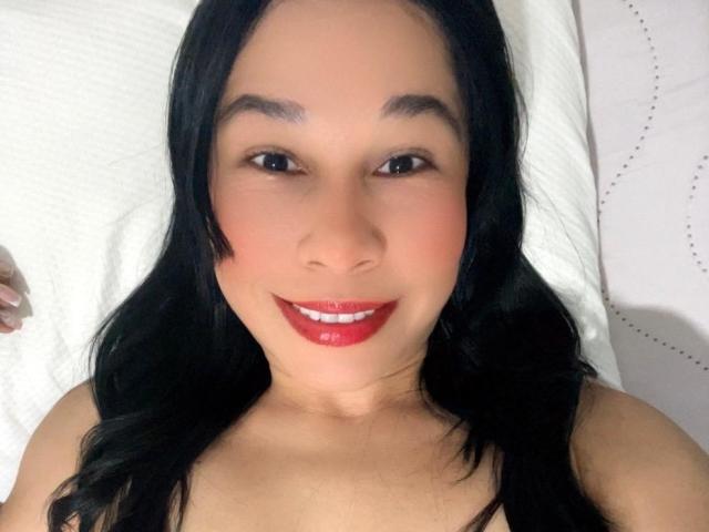 Nahaufnahme eines Fotos von IvannaX69 mit langen, schwarzen Haaren, heller Haut und rotem Lippenstift, die direkt in die Kamera lächelt. Sie hat dunkle Augenbrauen, eine sichtbare Ausschnitt und einen sexuellen, kokettierenden Ausdruck..