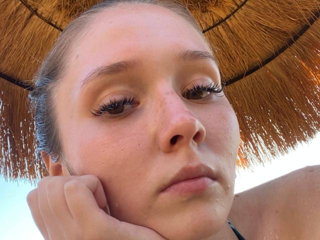 Close-up of MarniePlantier under a thatched umbrella, head resting on hand, eyes half-closed, wet skin, small droplets on face, natural light, subtle sexual tension, bare shoulders, neutral expression.