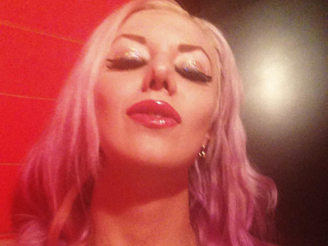 Close-up van RoseVampire met roze haar, veel make-up en glanzende rode lippen, ogen half gesloten, verlicht door fel licht. rode en zwarte achtergrond, intense blik, lichte onscherpte, uitdagende uitdrukking.