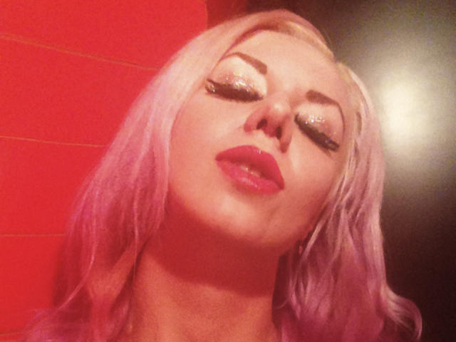 Nahaufnahme eines Fotos von RoseVampire mit platinblondem Haar, starkem Make-up und glänzenden roten Lippen. Augen geschlossen, Lippen leicht geöffnet. Helles, leuchtendes Rot als Hintergrund mit einer hellen Lichtquelle. Verdammt heiß..