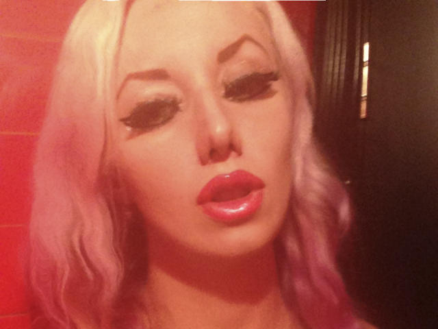 Photo en gros plan de RoseVampire, avec des cheveux blonds platine, un maquillage prononcé et des lèvres rouges brillantes. Les yeux à moitié fermés, elle dégage une tension sexuelle. Une lumière rouge projette un éclat séduisant sur son visage..