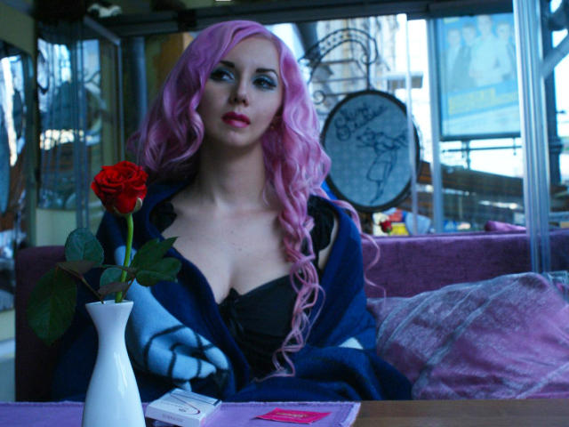 RoseVampire com cabelo rosa encaracolado, vestindo um vestido preto e jaqueta azul, sentada em um café, segurando uma rosa vermelha, sexy. ambiente, mesa com cartas e vaso, iluminação fraca.