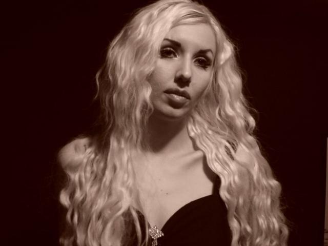 Schwarz-weißes Foto von RoseVampire mit langen, welligen, platinblonden Haaren, intensivem Blick und einem sexy, selbstbewussten Ausdruck. Ausdruck. Sie trägt ein schwarzes Oberteil mit tiefem Ausschnitt und eine Anhänger-Kette..