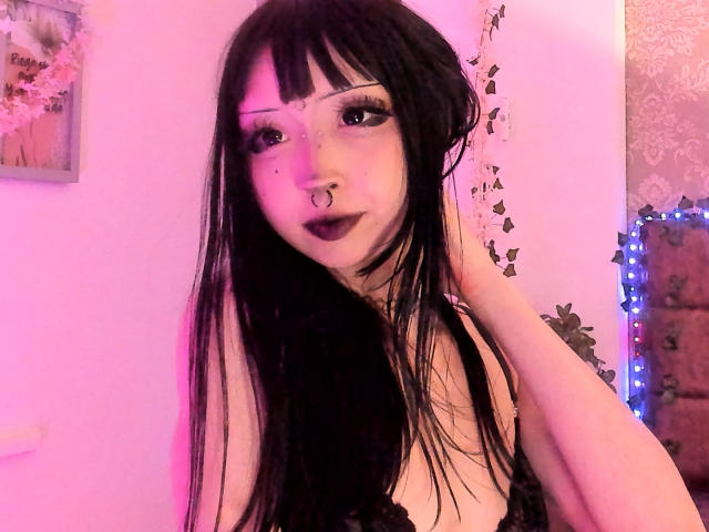 MillaHottLove69 hat langes, schwarzes Haar, blasse Haut und dunklen Lippenstift. Sie trägt eine schwarze Spitzenbluse, blickt direkt in die Kamera und lächelt leicht, ihre Hand ist in der Nähe ihres Gesichts. Rosa und violette Beleuchtung.