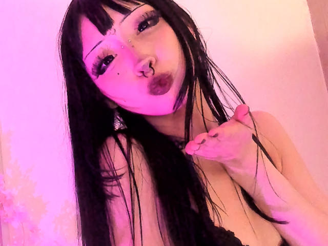 Close-up foto van MillaHottLove69, een vrouw met bleke huid, lang zwart haar, veel make-up en een neusring. Ze steekt haar tong uit, met haar hand in de buurt van haar sleutelbeen. Roze en paarse verlichting.