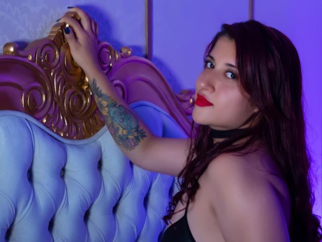 Alis Roberts, met donkerbruin haar en rode lippenstift, leunt tegen een blauw gestoffeerd hoofdeinde en houdt een sierlijk houten frame vast. met haar getatoeëerde arm. Blauwe en paarse verlichting versterken de sfeer..