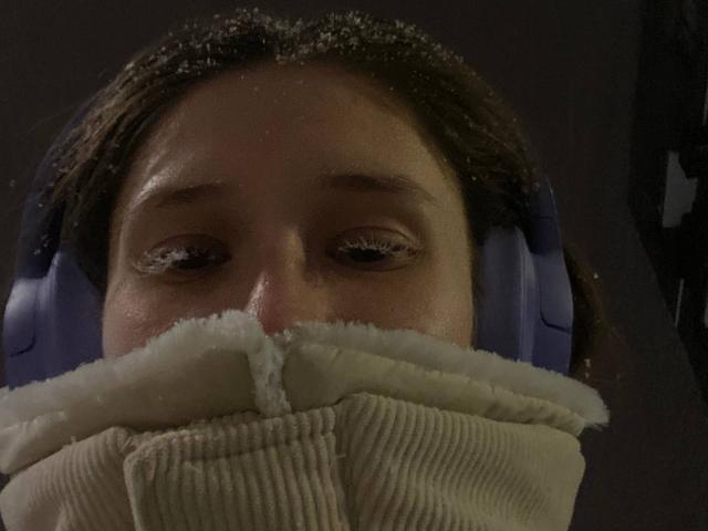 Close-up of MarniePlantier's face with a thick, beige scarf covering her mouth and nose. She wears blue headphones, gazing forward with slightly tired eyes. Dark, shadowy background. Sexual tension.