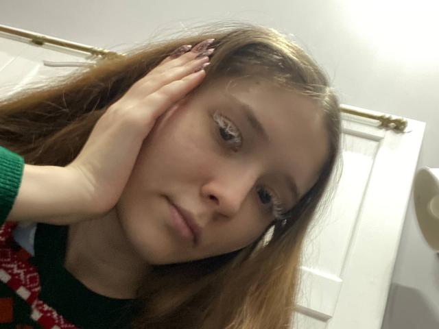 Close-up van MarniePlantier, met bruin haar, hand op haar wang, serieuze uitdrukking, draagt een groene en rode trui, helder. Badkamervlamp achter, schaduwen op het gezicht, lichtelijk rommelig haar, zachte focus, casual selfie-gevoel..