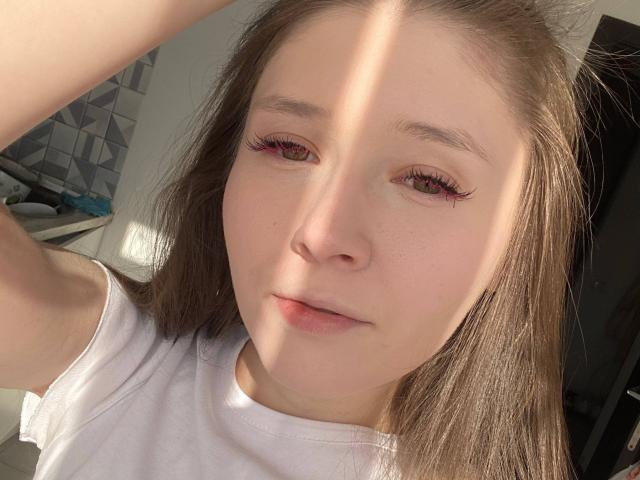 Close-up of MarniePlantier, light-skinned with brown hair, wearing a white shirt. Sunlight casts diagonal shadows across her face, highlighting her freckles and subtle, half-lidded expression. Fucking gorgeous.