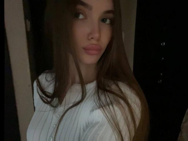 Fotografía de Emerald Weinreb, una mujer atractiva con cabello castaño largo, ojos verdes y labios rosados y carnosos, vestida con un ajustado vestido blanco. Una camisa que realza sus grandes senos, mirando hacia un lado..