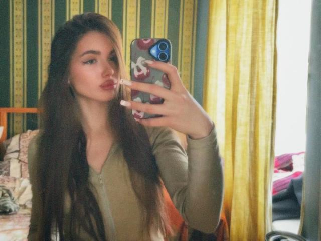 Fotografia de Emerald Weinreb, tirando uma selfie no espelho com um smartphone. Ela tem cabelos castanhos longos, lábios cheios e um moletom verde com zíper. Cortinas amarelas e papel de parede listrado ao fundo. Incrivelmente sexy..