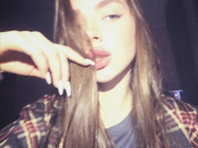 Blurred photo of EmeraldWeinreb, with long brown hair, licking a finger with long white nails. Wearing a plaid shirt over a gray top. Background is dark. Slight sexual tension.