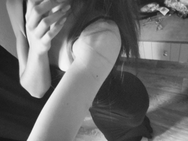 Black and white photograph of EmeraldWeinreb, close-up of her arm and hand, slight shadow on her upper thigh, partial view of her face, blurry background, sensual and intimate atmosphere.