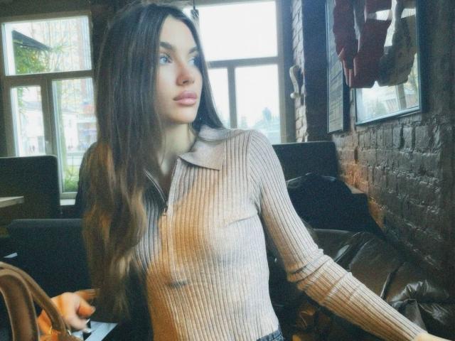 Fotografía de Emerald Weinreb, una joven con cabello largo y castaño, vestida con una blusa beige transparente y con textura, sentada en una cafetería con poca luz, con paredes de ladrillo. Su expresión es sensual, con una tensión sexual evidente..