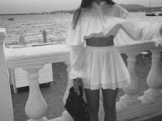 Black-and-white photo of EmeraldWeinreb standing by a railing, wearing a white, flowing dress with a wide belt, holding a bag, with a body of water and distant lights in the background.