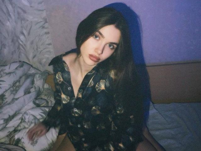 Photographie d'Emerald Weinreb, assise sur un lit, portant une chemise noire ornée de motifs en feuille d'or. Elle a de longs cheveux noirs, une peau pâle et des yeux bleus perçants, avec une tête légèrement inclinée, d'une manière séduisante..