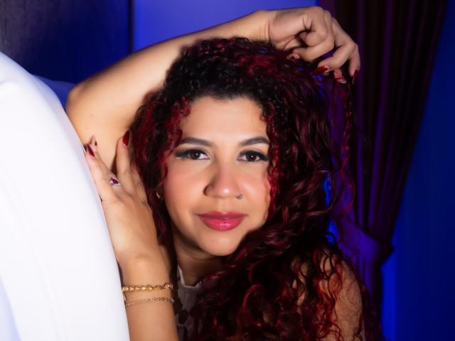 Antonela Riveiro mit lockigem, rotem Haar, Nasenring und rotem Lippenstift, die ihre Haare hält, lächelnd, trägt ein goldenes Armband, ein weißes Hemd, und im Hintergrund sind dunkelrote Vorhänge zu sehen..