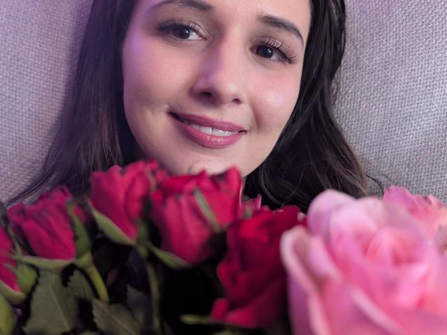 Fotografía de primer plano de Jenna-sxy19-hot, con piel clara y cabello castaño oscuro, sonriendo con labios rosados. Ella sostiene rosas rojas y rosas, creando un ambiente sensual con un aspecto suave y acogedor..