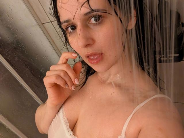 Fotografía de primer plano de Jenna-sxy19, con el pelo oscuro, húmeda, vistiendo un sujetador blanco de encaje, mirando seductoramente a la cámara. Gotas de agua en su piel, la mano derecha cerca de su boca..