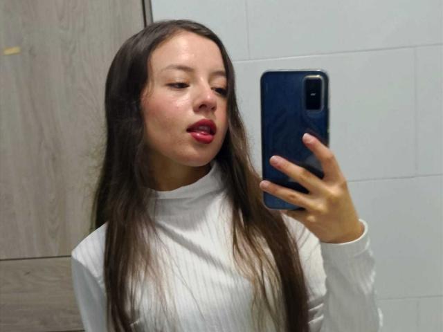 VioletaNova with long dark hair, red lipstick, and white turtleneck, holding a black phone in front of her face, slightly parted lips, sexual tension, casual selfie, wood and white tiled background.