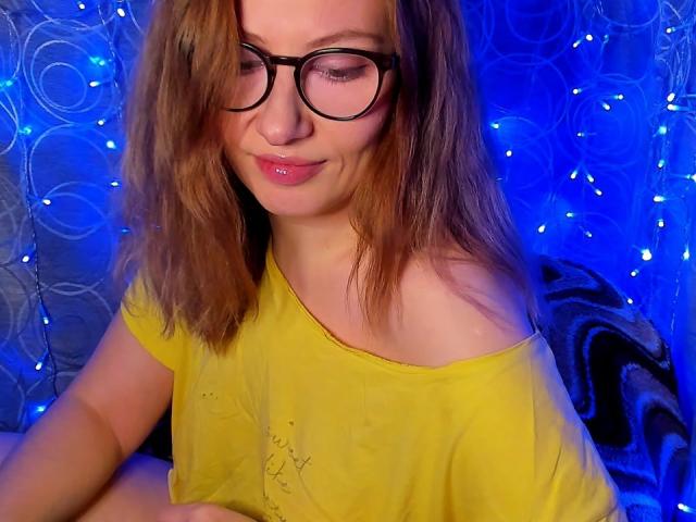 PaulinaSunny, trägt ein gelbes Top mit einem Schulterauschnitt, große, schwarz umrandete Brillen, braunes, schulterlanges Haar und rosa lippenstift, vor einem blauen Hintergrund mit kreisförmigen Mustern und Lichterketten. Total süß..