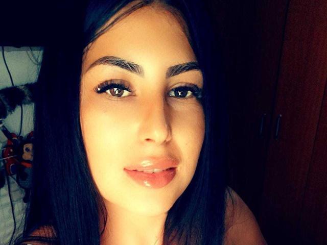 Close-up de IslaNoir, uma jovem com cabelos longos e pretos, olhos castanhos e lábios cheios, olhando diretamente para a câmera. Um leve sorriso. Iluminação forte, fundo escuro. Tensão sexual..