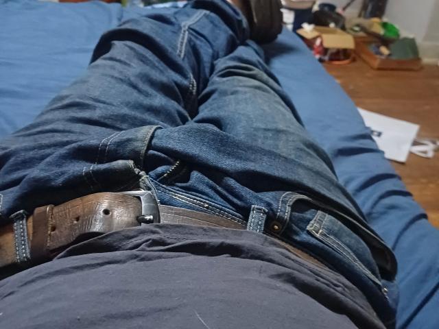 Close-up foto van het kruisgebied van maxKnd-hot, gekleed in strakke blauwe jeans met een bruine leren riem, liggend op een blauw bed. Gefocust op de uitstulping, die licht zichtbaar is door de denim..