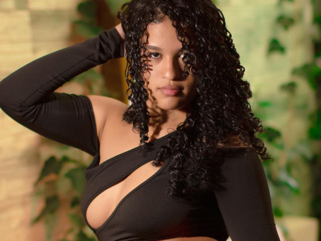 Photographie de Velaska Jones, avec des cheveux noirs et bouclés, portant un haut noir à une épaule avec un décolleté plongeant, qui expose sa décolleté. Elle lève la main gauche, et regarde directement l'appareil photo avec une expression séduisante..